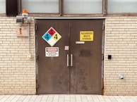 A metal door embedded in a brick wall with several warning signs. The signs include a hazardous materials placard with numbers, a yellow 'Hazardous Waste Storage' label, and a red 'Danger' notice warning against smoking and open flames. An orange warning light is mounted above, and there is a small vent on the wall.