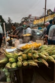 A street vendor stall is selling fresh corn, both raw and cooked. Steam is rising from a pot, and a variety of corn cobs are piled up on a table. Several people are standing around, some apparently purchasing and preparing food. The background includes other stalls, a car, and festively lit buildings, suggesting a busy market atmosphere.