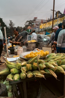 A street vendor stall is selling fresh corn, both raw and cooked. Steam is rising from a pot, and a variety of corn cobs are piled up on a table. Several people are standing around, some apparently purchasing and preparing food. The background includes other stalls, a car, and festively lit buildings, suggesting a busy market atmosphere.