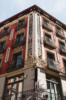 A historic corner building with ornate decorations and wrought iron balconies. The facade features intricate floral patterns and vibrant red and cream walls. There is signage reading 'Posada del Peine' above the corner, indicating the building's historical significance.