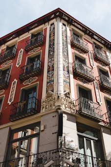 A historic corner building with ornate decorations and wrought iron balconies. The facade features intricate floral patterns and vibrant red and cream walls. There is signage reading 'Posada del Peine' above the corner, indicating the building's historical significance.