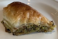 A piece of baklava with flaky, golden-brown phyllo dough layered on top of a dense filling that appears to contain crushed pistachios. The dessert has a shiny, syrupy finish, suggesting it is freshly prepared.