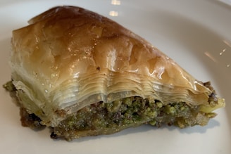 A piece of baklava with flaky, golden-brown phyllo dough layered on top of a dense filling that appears to contain crushed pistachios. The dessert has a shiny, syrupy finish, suggesting it is freshly prepared.