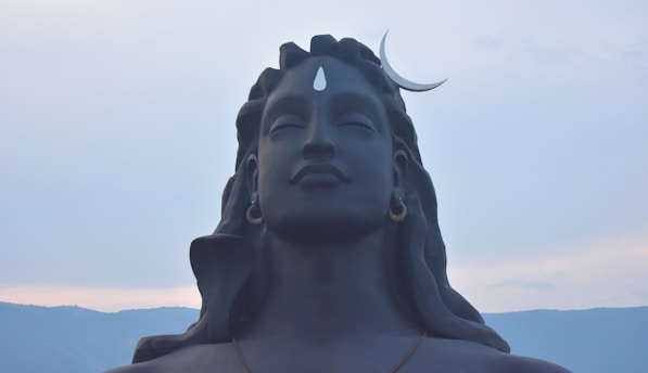 A large, serene statue of a figure with closed eyes and long hair. The sculpture features a crescent moon on the head and a prominent third eye on the forehead. The background consists of a light, cloudy sky with distant mountains.
