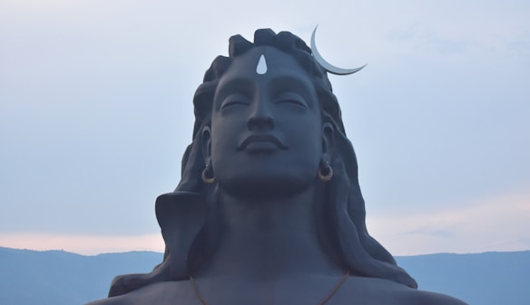 A large, serene statue of a figure with closed eyes and long hair. The sculpture features a crescent moon on the head and a prominent third eye on the forehead. The background consists of a light, cloudy sky with distant mountains.