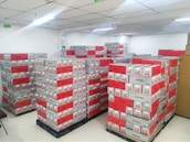 A room filled with stacked cardboard boxes organized on pallets. The boxes are labeled with the brand name Jozama and are predominantly red and gray. The room appears to be well-lit with bright overhead fluorescent lighting and has a clean, white tiled floor. There are shelves or cabinets on the right side of the image.