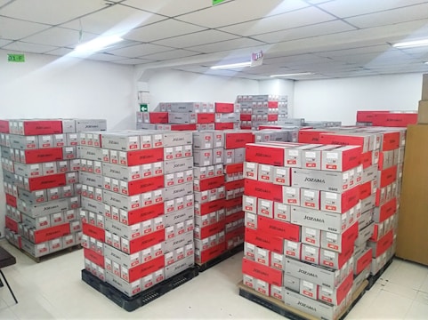 A room filled with stacked cardboard boxes organized on pallets. The boxes are labeled with the brand name Jozama and are predominantly red and gray. The room appears to be well-lit with bright overhead fluorescent lighting and has a clean, white tiled floor. There are shelves or cabinets on the right side of the image.