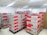 A room filled with stacked cardboard boxes organized on pallets. The boxes are labeled with the brand name Jozama and are predominantly red and gray. The room appears to be well-lit with bright overhead fluorescent lighting and has a clean, white tiled floor. There are shelves or cabinets on the right side of the image.