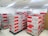 A room filled with stacked cardboard boxes organized on pallets. The boxes are labeled with the brand name Jozama and are predominantly red and gray. The room appears to be well-lit with bright overhead fluorescent lighting and has a clean, white tiled floor. There are shelves or cabinets on the right side of the image.