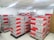 A room filled with stacked cardboard boxes organized on pallets. The boxes are labeled with the brand name Jozama and are predominantly red and gray. The room appears to be well-lit with bright overhead fluorescent lighting and has a clean, white tiled floor. There are shelves or cabinets on the right side of the image.