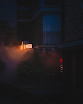 An atmospheric urban scene featuring a dimly lit exterior with a neon-lit sign partially shrouded in fog or smoke. The setting appears to be an old building with visible balconies and a moody, mysterious ambiance. Soft red and orange lights add a warm glow to the predominantly dark environment.