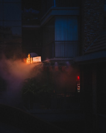 An atmospheric urban scene featuring a dimly lit exterior with a neon-lit sign partially shrouded in fog or smoke. The setting appears to be an old building with visible balconies and a moody, mysterious ambiance. Soft red and orange lights add a warm glow to the predominantly dark environment.