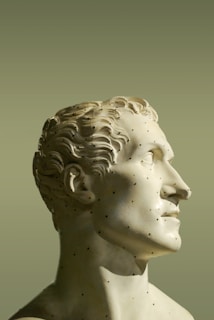 A classical marble statue of a man with a detailed and serene expression. His head is slightly tilted upward, and the sculpture captures intricate features such as curly hair and a smooth surface. The background is a muted grayish-green color.
