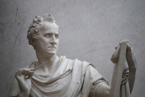 A detailed sculpture of a man holding a chisel and partially draped in cloth. The figure's expression is contemplative, with curly hair and classical attire, gazing intently forward.