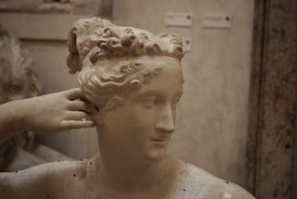 A classical marble statue of a woman's head and upper body, showcasing intricate hair details and a serene expression. The statue's hand is raised to the side of the head, touching the ear. The setting appears to be an indoor exhibition space.