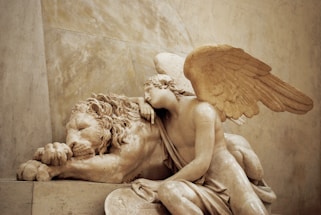 A detailed stone sculpture depicts a winged figure leaning gently over a resting lion. The lion appears to be asleep, with its head resting on its paws. The winged figure, resembling an angel, has a contemplative expression and drapes an arm over the lion, creating a sense of companionship and peace.