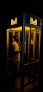 A person is standing inside an illuminated phone booth at night, speaking into a telephone. The scene is dimly lit with the glow from the booth's interior light providing the main source of illumination against the dark background.