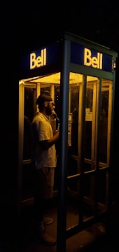 A person is standing inside an illuminated phone booth at night, speaking into a telephone. The scene is dimly lit with the glow from the booth's interior light providing the main source of illumination against the dark background.