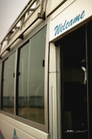 A close-up view of a vehicle, possibly a van or bus, featuring a metallic exterior with large glass windows. The word 'Welcome' is visibly written in blue script on the side near the entrance. The image gives a partial glimpse of the interior through an open door.