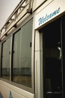 A close-up view of a vehicle, possibly a van or bus, featuring a metallic exterior with large glass windows. The word 'Welcome' is visibly written in blue script on the side near the entrance. The image gives a partial glimpse of the interior through an open door.
