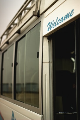 A close-up view of a vehicle, possibly a van or bus, featuring a metallic exterior with large glass windows. The word 'Welcome' is visibly written in blue script on the side near the entrance. The image gives a partial glimpse of the interior through an open door.