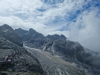 A rugged mountain landscape with steep gray rock formations and a cloudy sky. A pathway with several people in colorful clothing is visible, winding its way up the mountain slope. Mist rolls through the scene, giving it an ethereal quality.