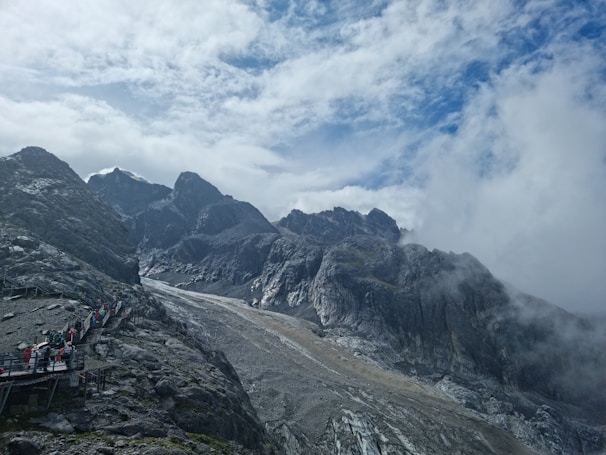 A rugged mountain landscape with steep gray rock formations and a cloudy sky. A pathway with several people in colorful clothing is visible, winding its way up the mountain slope. Mist rolls through the scene, giving it an ethereal quality.