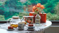 A table is set with a teapot filled with tea, a patterned cup on a saucer, and several mooncakes on white plates. A colorful, vintage-style tin sits next to a glass vase containing orange flowers. The table is covered with a cloth and positioned in front of a large window overlooking lush greenery.