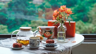 A table is set with a teapot filled with tea, a patterned cup on a saucer, and several mooncakes on white plates. A colorful, vintage-style tin sits next to a glass vase containing orange flowers. The table is covered with a cloth and positioned in front of a large window overlooking lush greenery.