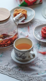 A wooden-lidded glass teapot with floral tea sits on a table adorned with a lace cloth. Nearby, a white teacup with a decorative pattern contains tea. A couple of plates hold mooncakes, positioned in a soft, inviting, afternoon light.