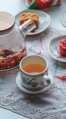 A wooden-lidded glass teapot with floral tea sits on a table adorned with a lace cloth. Nearby, a white teacup with a decorative pattern contains tea. A couple of plates hold mooncakes, positioned in a soft, inviting, afternoon light.