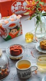 An arrangement featuring a colorful tea set with cups filled with tea, and a glass jar with floral designs. Mooncakes shaped like lions are placed on plates, and there is a decorative box with a cartoon character in a traditional festival outfit. Red flowers in a glass vase enhance the festive atmosphere.