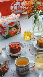 An arrangement featuring a colorful tea set with cups filled with tea, and a glass jar with floral designs. Mooncakes shaped like lions are placed on plates, and there is a decorative box with a cartoon character in a traditional festival outfit. Red flowers in a glass vase enhance the festive atmosphere.