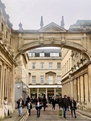 An architectural archway connects two classical buildings with ornate details. Below, a pedestrian street is busy with people walking, some carrying shopping bags. The architecture features columns and a stone facade.