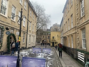 A narrow street lined with classical stone buildings, featuring an outdoor seating area with metallic tables and chairs. A few people walk along the cobblestone path, with one person in a red jacket. There's a sign for a Georgian tearoom, and the street is flanked by tall, bare-branched trees.