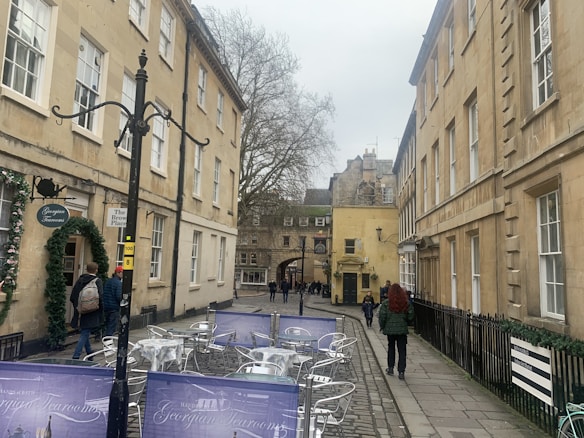 A narrow street lined with classical stone buildings, featuring an outdoor seating area with metallic tables and chairs. A few people walk along the cobblestone path, with one person in a red jacket. There's a sign for a Georgian tearoom, and the street is flanked by tall, bare-branched trees.