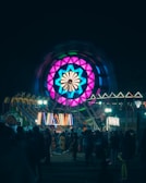 A vibrant carnival scene featuring a large Ferris wheel illuminated with colorful lights in pink, blue, and teal. The image is set at night, with various other illuminated attractions in the background and a crowd of people enjoying the festivities.