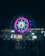 A vibrant carnival scene featuring a large Ferris wheel illuminated with colorful lights in pink, blue, and teal. The image is set at night, with various other illuminated attractions in the background and a crowd of people enjoying the festivities.