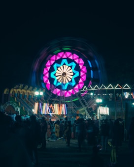 A vibrant carnival scene featuring a large Ferris wheel illuminated with colorful lights in pink, blue, and teal. The image is set at night, with various other illuminated attractions in the background and a crowd of people enjoying the festivities.