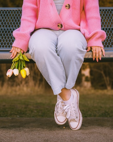 A person wearing a pink cardigan, light blue pants, and white sneakers is sitting on a bench. The person's hands are resting on the bench, and they are holding a small bouquet of tulips with white, pink, and yellow petals. The background is slightly blurred and appears to be an outdoor setting.