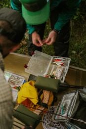 Two people are organizing fishing gear outdoors. Various fishing lures and equipment are spread out on a surface, including a yellow backpack, a box of colorful lures, and other tackle. One person is handling small fishing components.