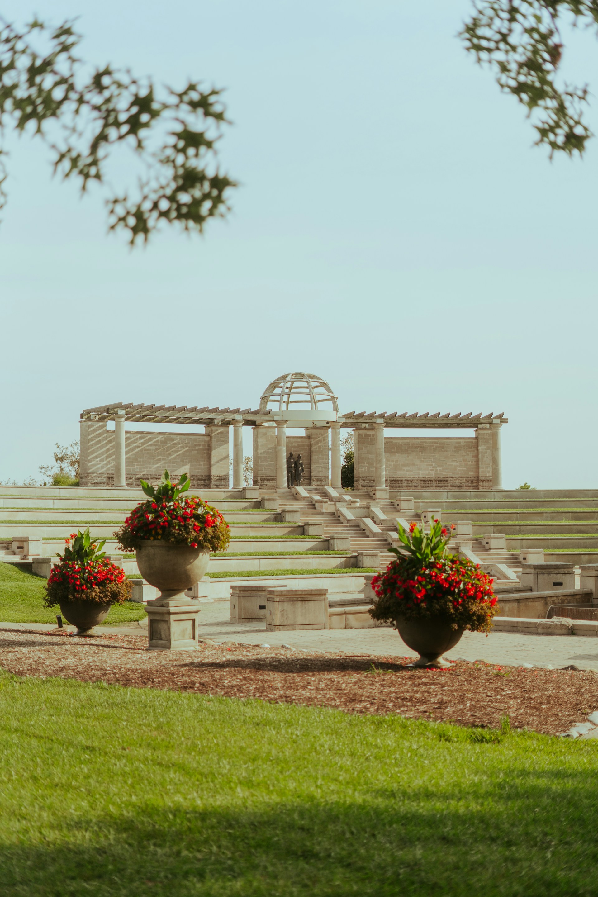 A landscaped garden featuring large stone planters filled with red and green plants is in the foreground. Beyond, a tiered stone amphitheater with steps leads up to a stone structure with columns and a domed roof. The ground is covered in green grass, and there are leafy branches in the upper corners.