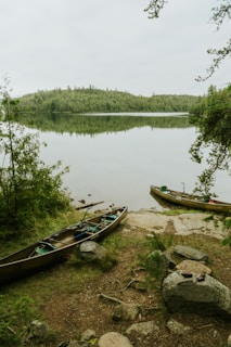 A serene lakeside scene with two canoes resting on the rocky shore. The calm water reflects the surrounding dense forest, creating a peaceful and natural atmosphere. The sky is overcast, adding a soft, muted light to the surroundings.
