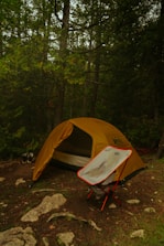 A yellow camping tent is set up in a forested area with dense green foliage and tall trees surrounding it. In the foreground, a foldable camping chair with red legs is placed in front of the tent. The ground is covered with patches of grass, dirt, and scattered rocks. A pair of hiking shoes is visible beside the tent.