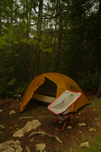A yellow camping tent is set up in a forested area with dense green foliage and tall trees surrounding it. In the foreground, a foldable camping chair with red legs is placed in front of the tent. The ground is covered with patches of grass, dirt, and scattered rocks. A pair of hiking shoes is visible beside the tent.