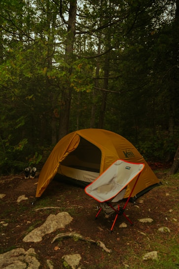 A yellow camping tent is set up in a forested area with dense green foliage and tall trees surrounding it. In the foreground, a foldable camping chair with red legs is placed in front of the tent. The ground is covered with patches of grass, dirt, and scattered rocks. A pair of hiking shoes is visible beside the tent.