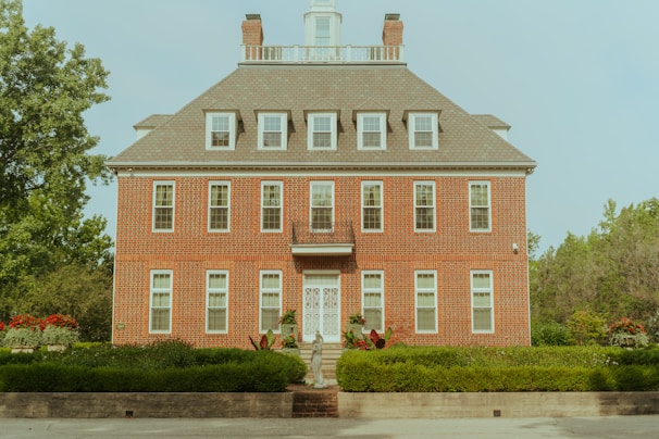 A symmetrical three-story brick building with a steeply pitched roof and multiple windows. The entrance features white double doors, and a set of steps leads up to it. Surrounding the building are well-maintained gardens, with green bushes and some red flowers visible. Trees are present in the background, contributing to a peaceful suburban setting.