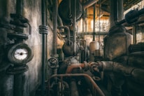 A complex network of industrial pipes and valves fills the space, with aged metal surfaces showing signs of rust and wear. A pressure gauge is prominently visible, surrounded by various cylindrical tanks and intricate piping connected throughout the facility. The lighting casts a warm glow, highlighting the dusty and gritty atmosphere typical of an abandoned or old industrial site.