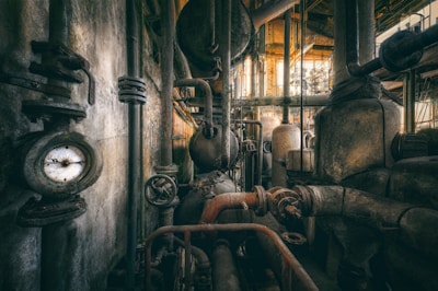 A complex network of industrial pipes and valves fills the space, with aged metal surfaces showing signs of rust and wear. A pressure gauge is prominently visible, surrounded by various cylindrical tanks and intricate piping connected throughout the facility. The lighting casts a warm glow, highlighting the dusty and gritty atmosphere typical of an abandoned or old industrial site.