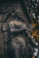A detailed angelic sculpture stands against a background of intricate architectural designs, with weathered textures and a somber expression. The figure has wings and its arms are crossed over its chest. The surroundings include hints of foliage with autumn colors, adding depth and contrast to the stonework.